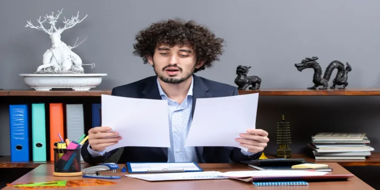 A young professional reviewing CV personal statement drafts at his office desk, surrounded by documents and folders, representing the process of writing a good CV personal statement.